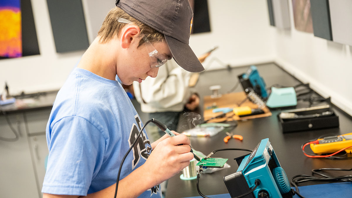A boy in safety glasses solders a circuit board in a classroom lab.