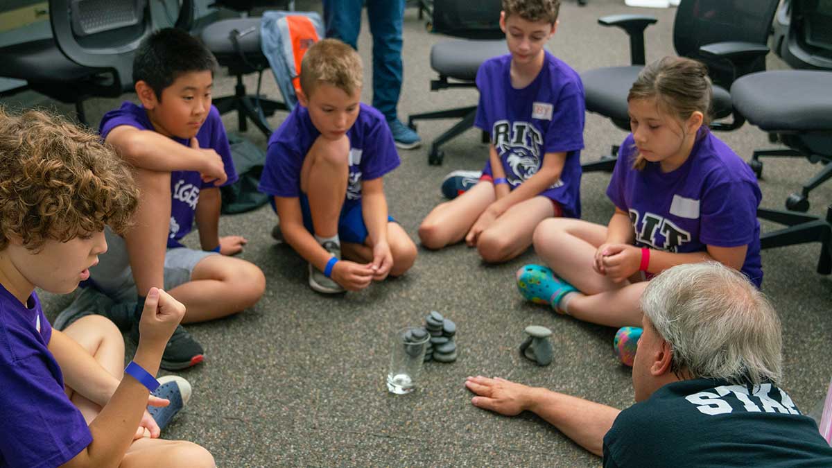 Children in purple shirts sit on the floor with an instructor, stacking rocks in a group activity.