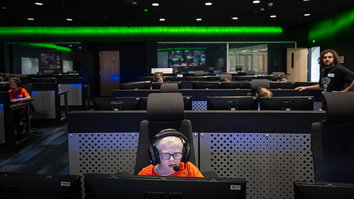 A child wearing headphones plays a computer game in a dark esports lab with multiple monitors.