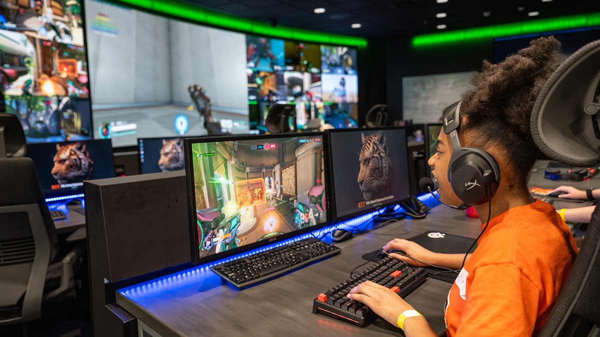 A girl with headphones plays a video game at a computer in a large cybersecurity lab with large screens.
