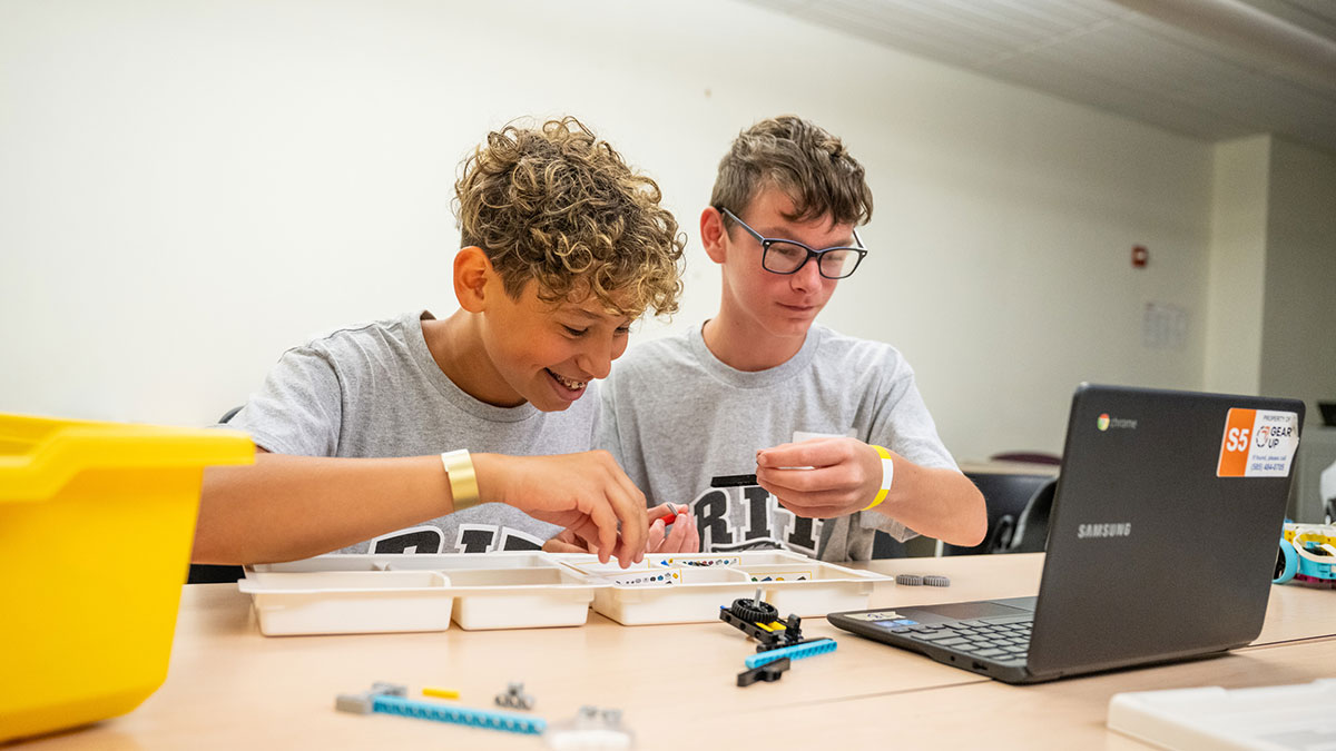 Two boys in matching shirts assemble LEGO robotics pieces while using a laptop.