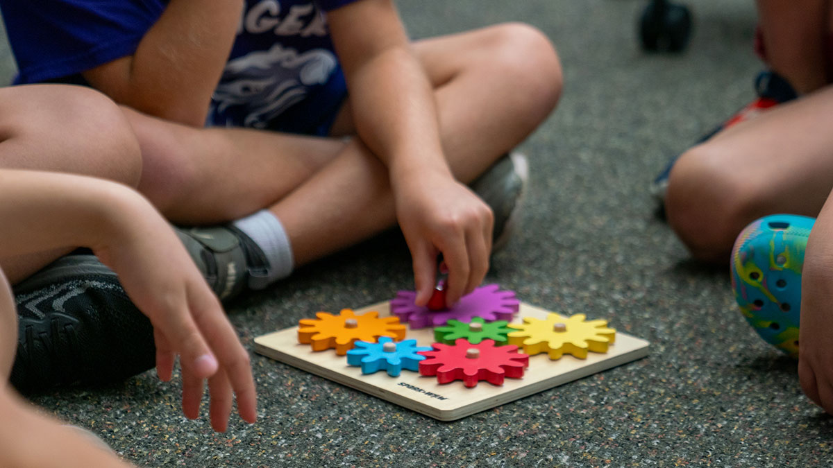 Children sit cross-legged on the floor, working together on a colorful wooden gear puzzle.
