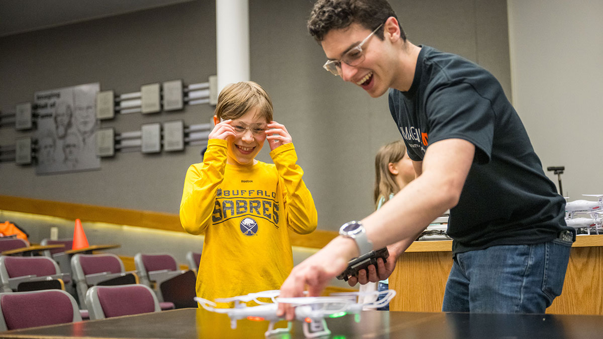 A boy in a yellow shirt smiles as an R I T student helps him operate a small drone in a classroom.