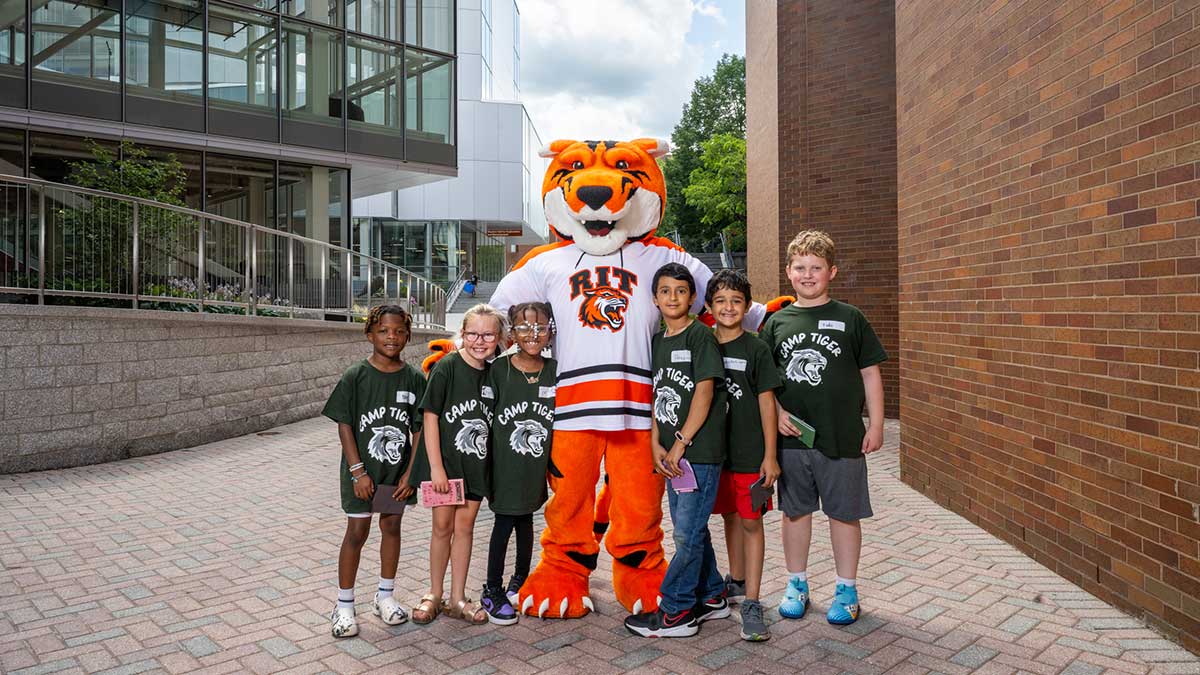 Children wearing Camp Tiger shirts pose outdoors with the R I T tiger mascot.