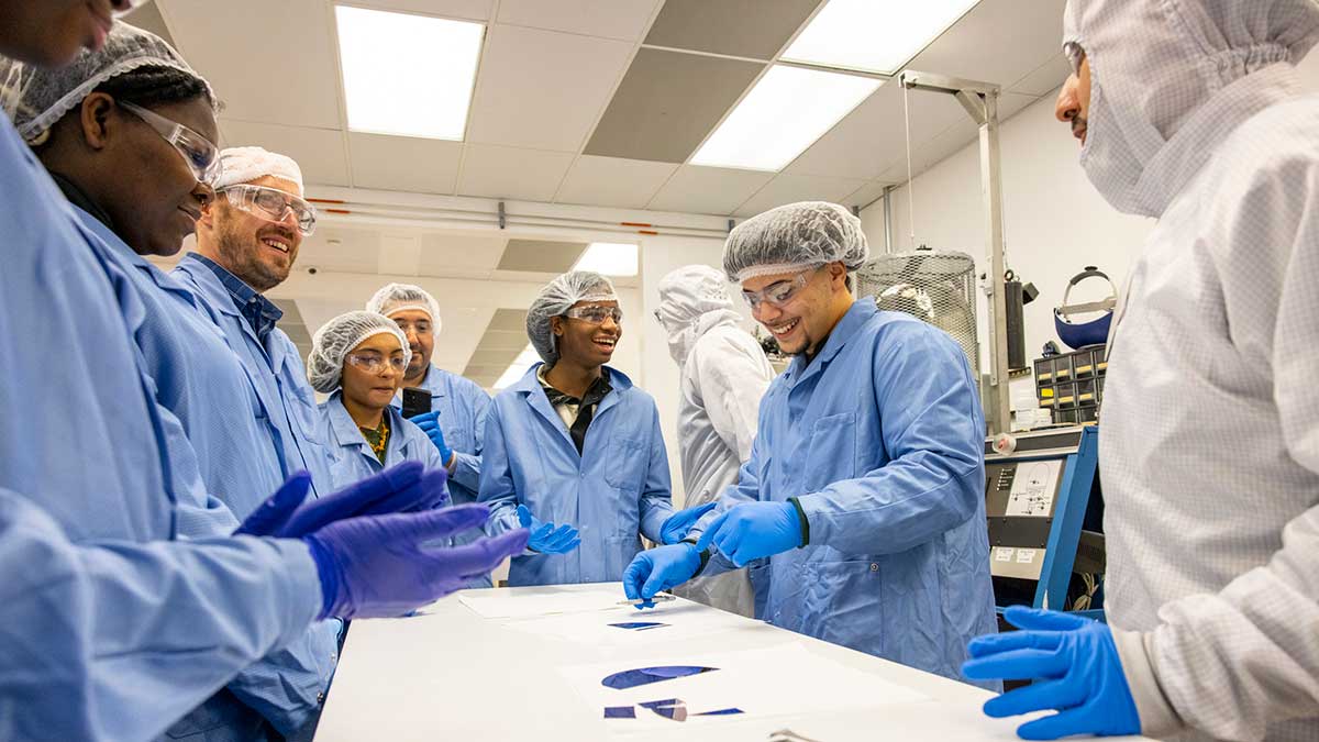 Students in protective blue lab coats and hairnets observe a demonstration inside a cleanroom.