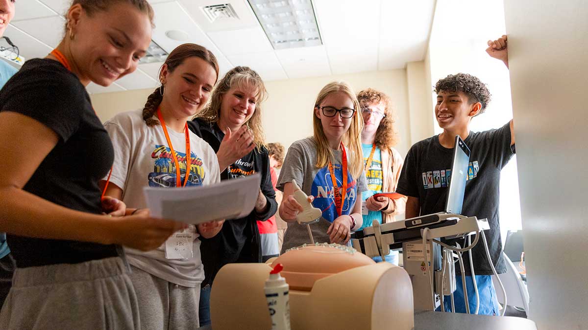 Teens in a classroom laugh and watch as one participant uses an ultrasound device on a training mannequin.