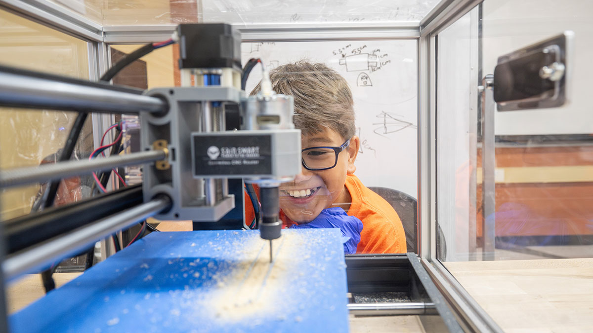 A child peers closely and smiles while operating a C N C milling machine through a protective enclosure.
