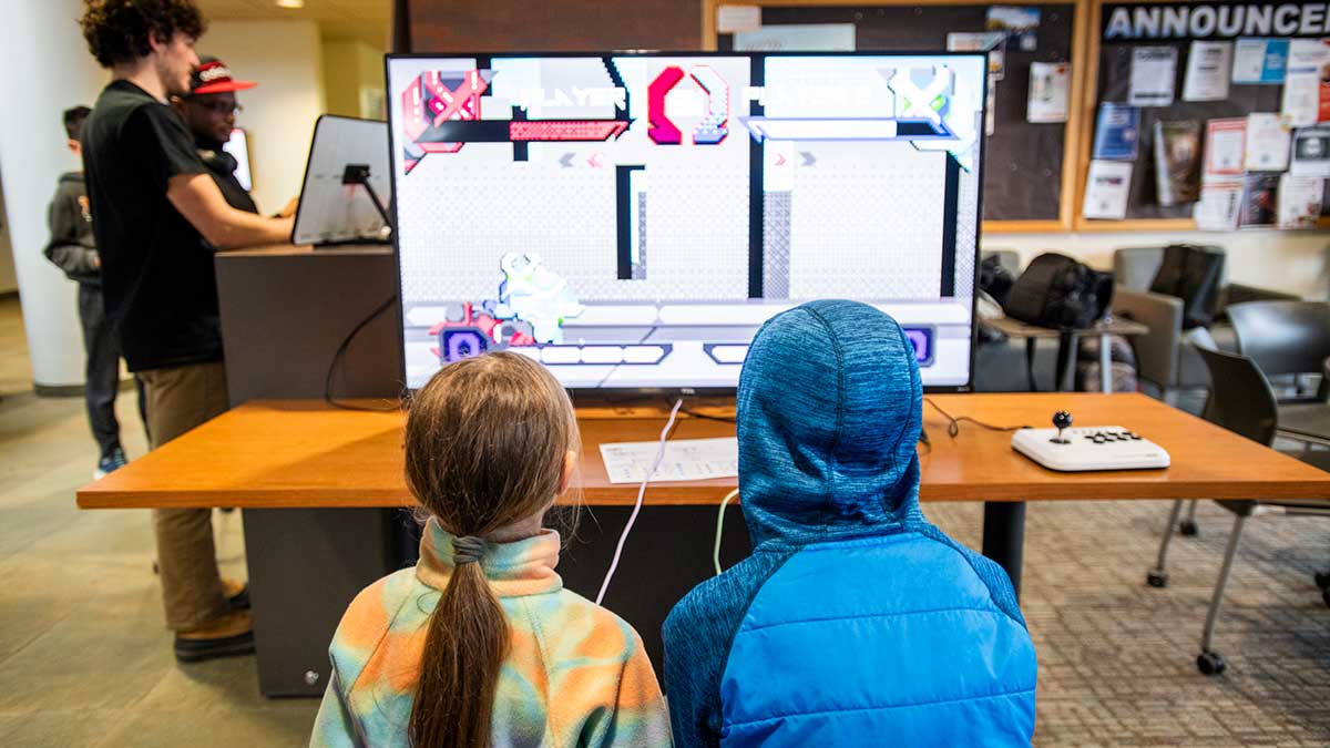 Two children sit facing a large monitor while playing a video game with an arcade-style joystick.
