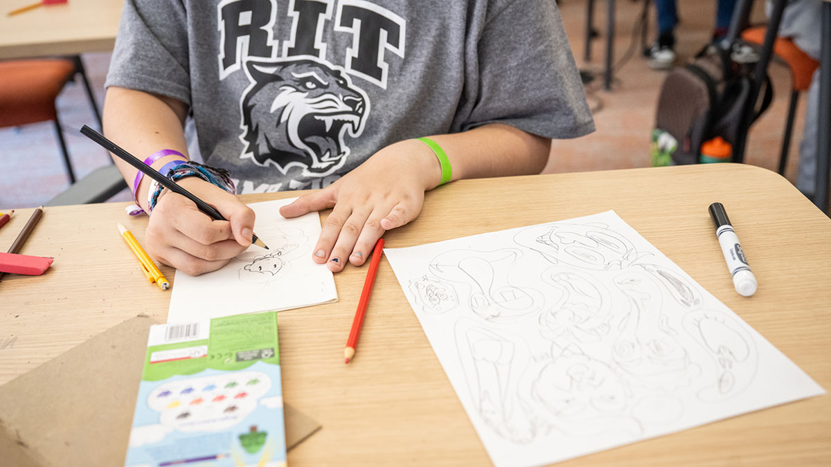 A child in an R I T t-shirt sketches cartoon characters with colored pencils at a classroom desk.