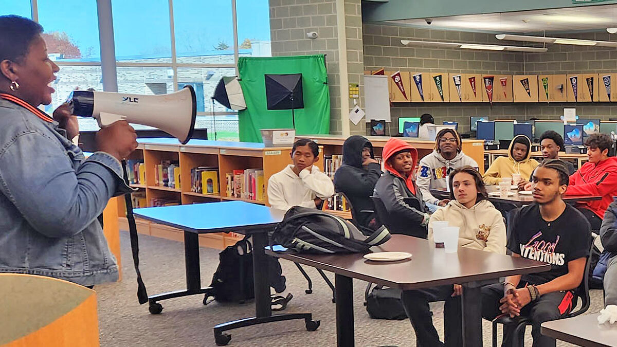 a woman uses a bullhorn to address a large classroom of high school students.