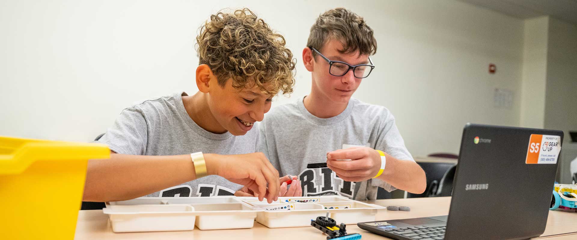 Two boys in matching shirts work together on a LEGO robotics kit beside a laptop.