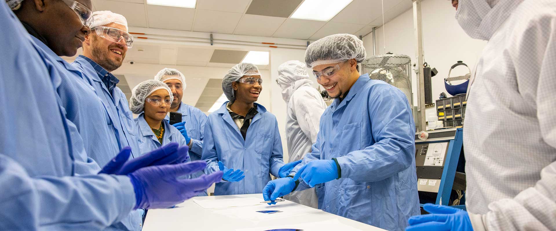 High school students in cleanroom suits and goggles smile while examining materials during a lab tour.