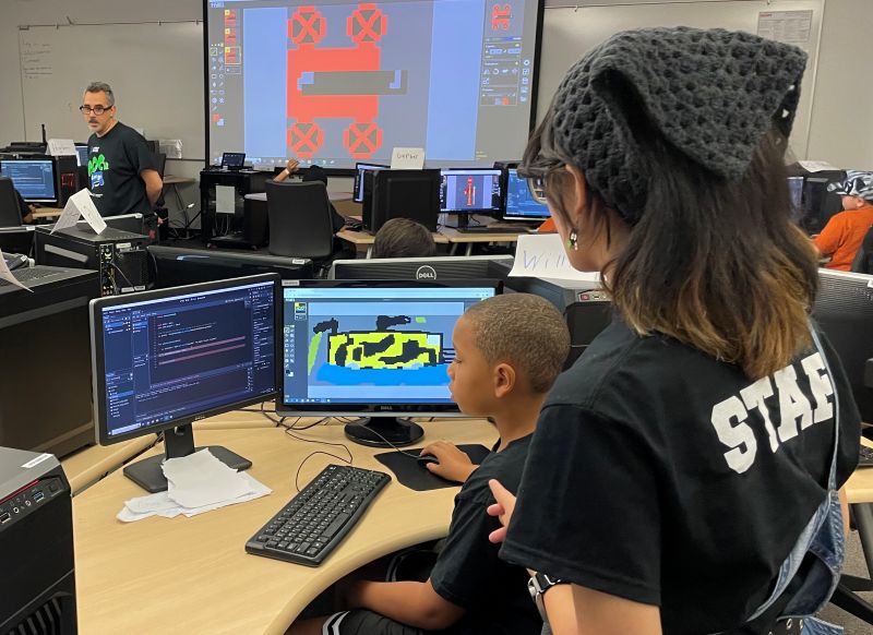 Staff member working with a boy at a computer, learning to create a video game in a computer lab.