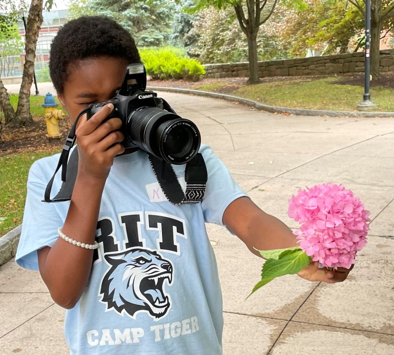 Girl taking a picture of a large pink flower with a camera outdoors.