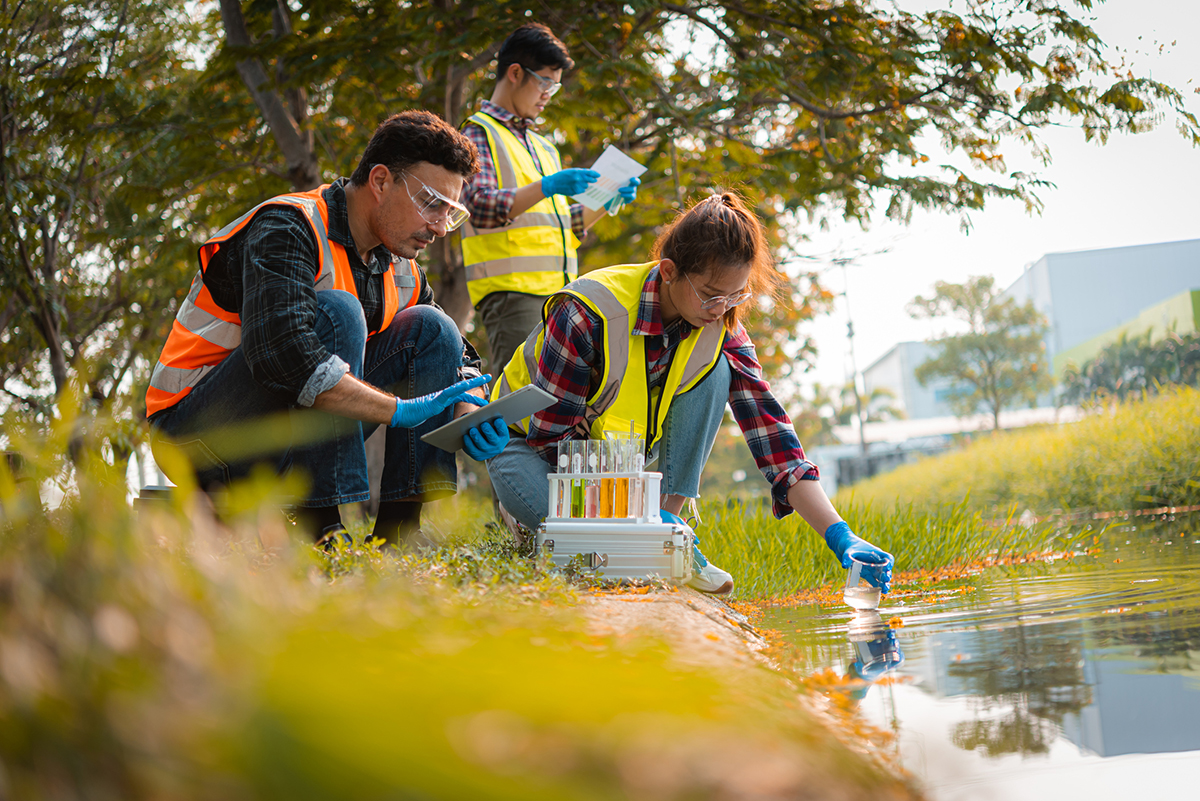 Three students collecting water samples from a pond outdoors.