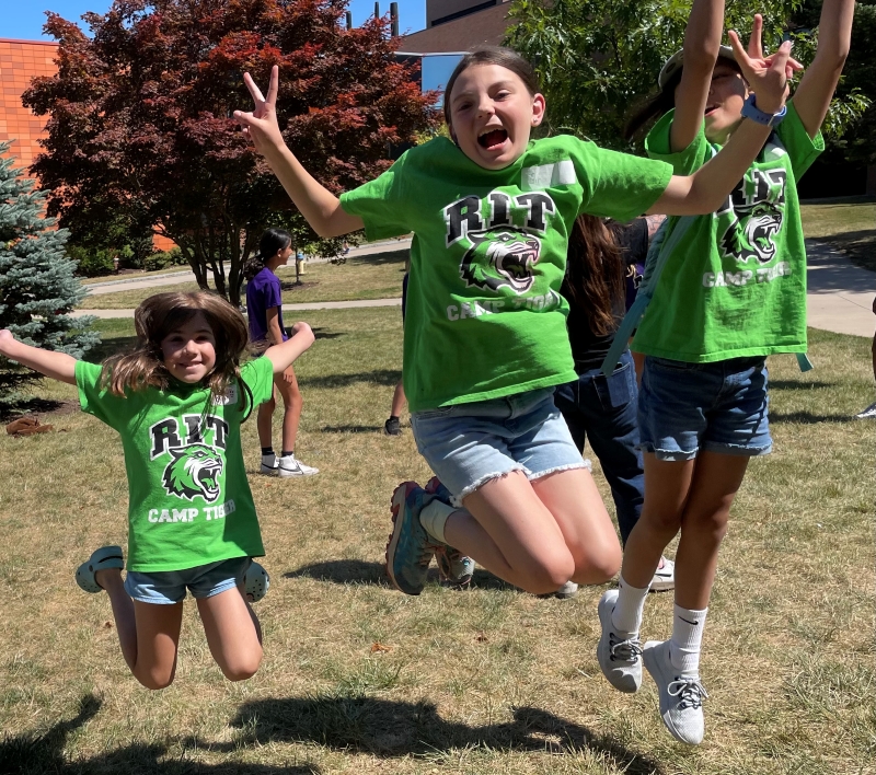 Three girls in green Camp Tiger t-shirts jumping outdoors on the grass.