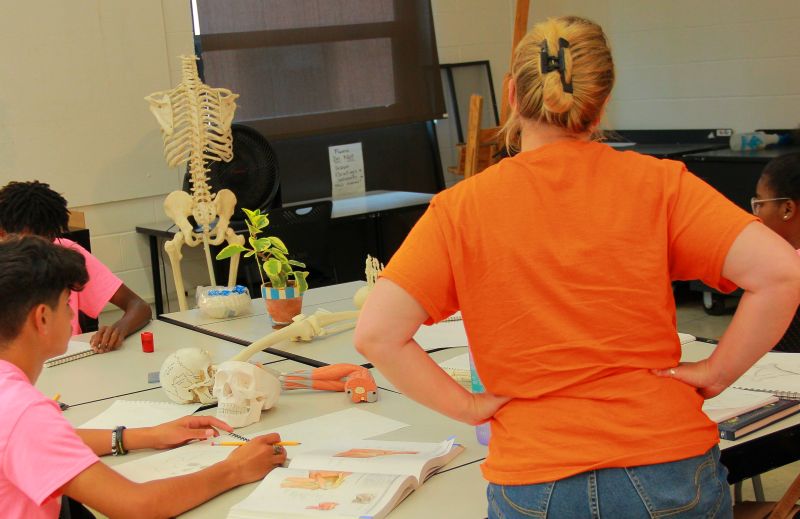 Three teens working with an instructor to draw human skeleton bones in a classromm.