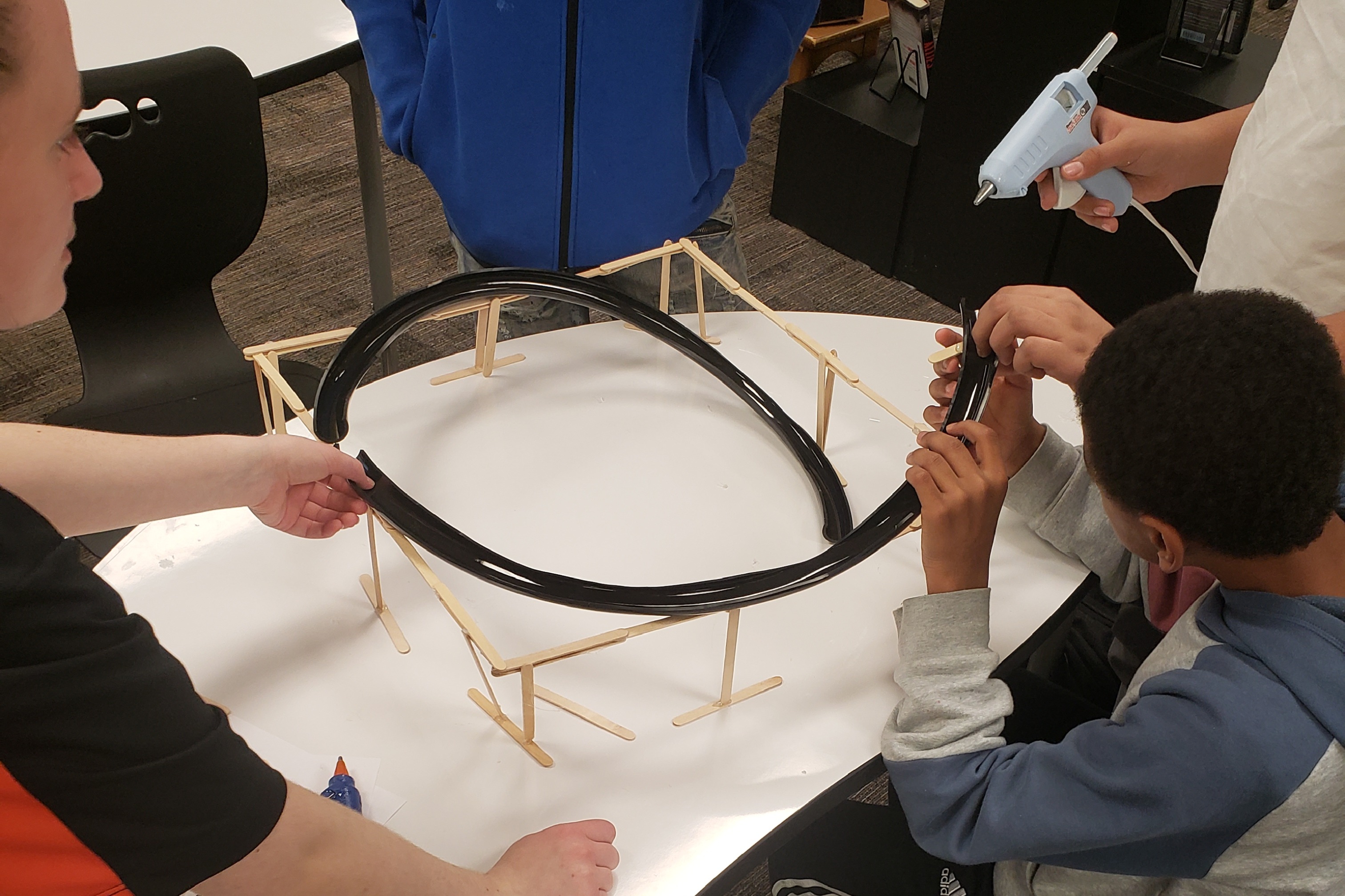 RIT student helping  outh create a theme park ride using glue gun and a variety of materials.