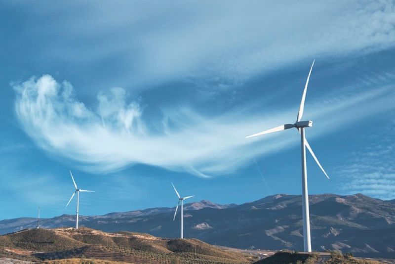 Outdoor picture of 3 wind turbines with mountains in the background and wispy clouds in the sky.