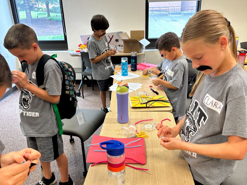 Several youth create villain disquises including mustaches in a classroom. 