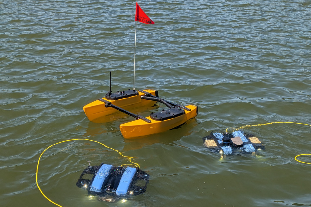 Yellow autonomous surface boat with a red flag floating on water alongside two blue underwater vehicles
