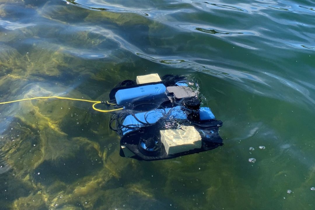 A small remotely operated underwater vehicle (ROV) floating near the surface of a clear, greenish lake, tethered by a yellow cable, with a forested shoreline in the background.