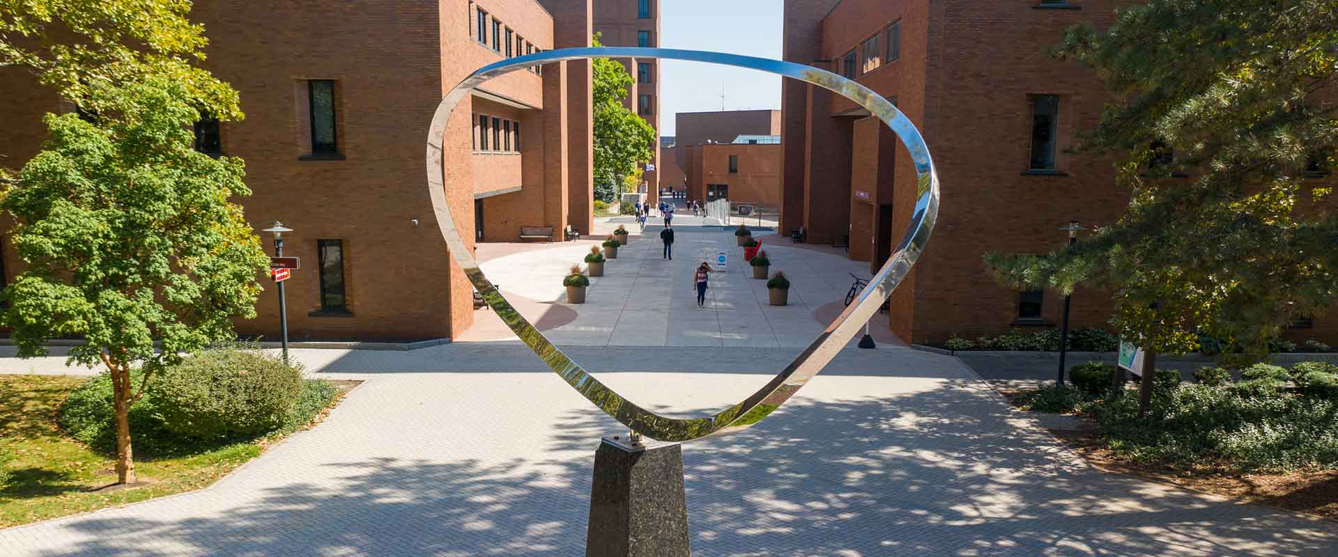 a view of two brick buildings separated by a brick walkway, as seen through the metal loop of a statue.