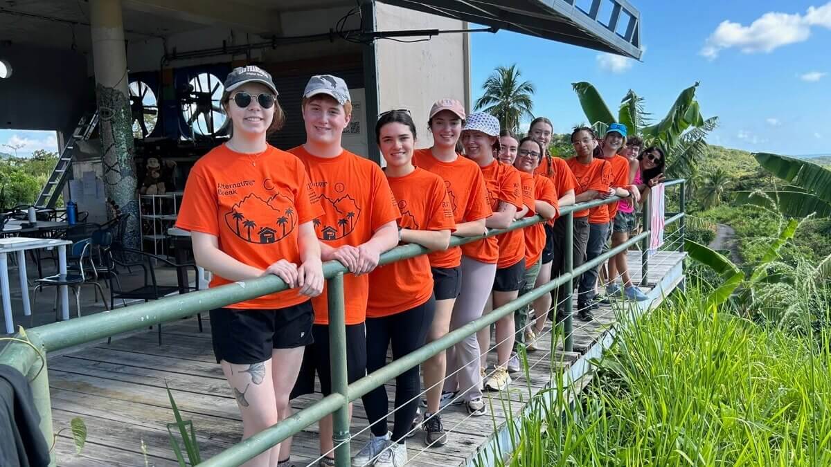 A group of students during Alternative Break posing for a picture in a line outside on a balcony in a grassy location