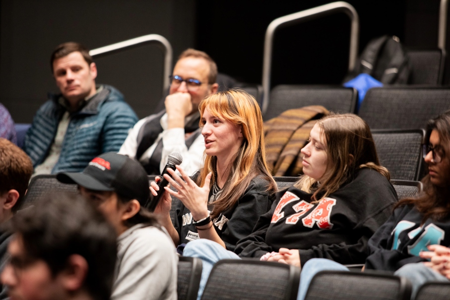 A young woman with orange-streaked hair holds a microphone and speaks while sitting in a dark lecture hall or auditorium with several other attendees.