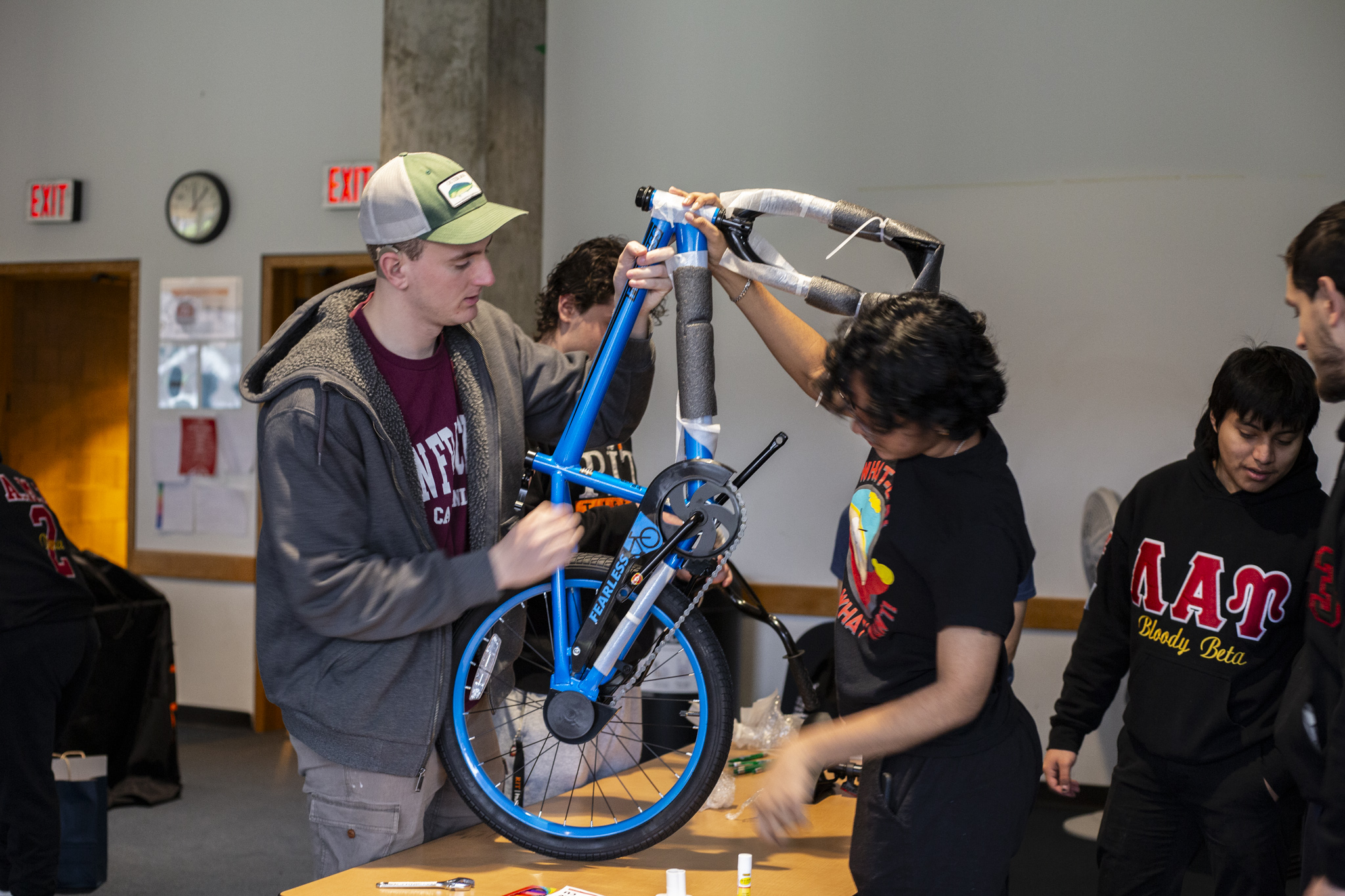 Two people work together to assemble a blue bicycle on a table indoors, with tools and bike parts visible around them.