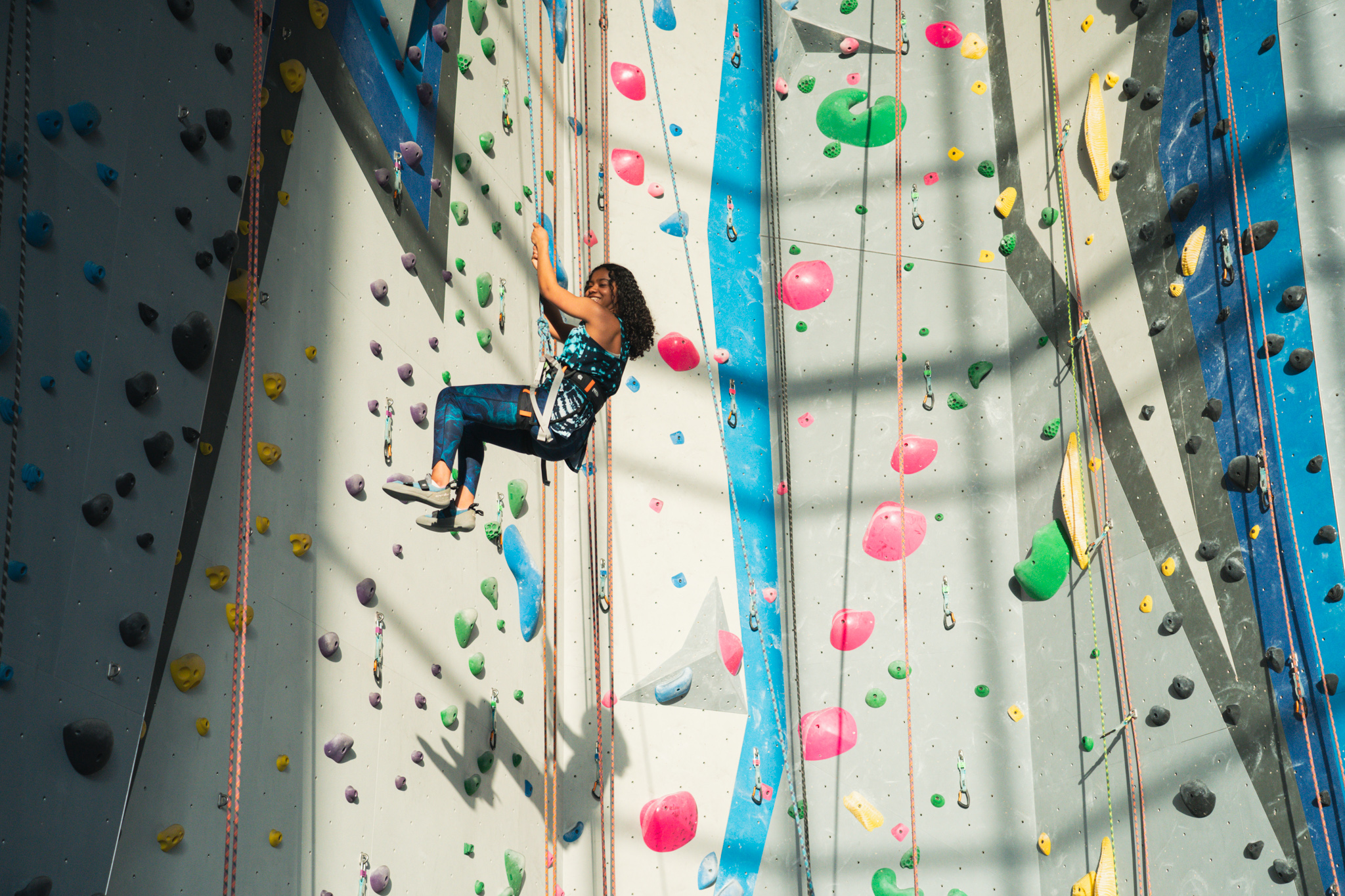 Person secured with a harness climbs an indoor rock wall covered in colorful holds and ropes, with sunlight casting shadows across the surface.