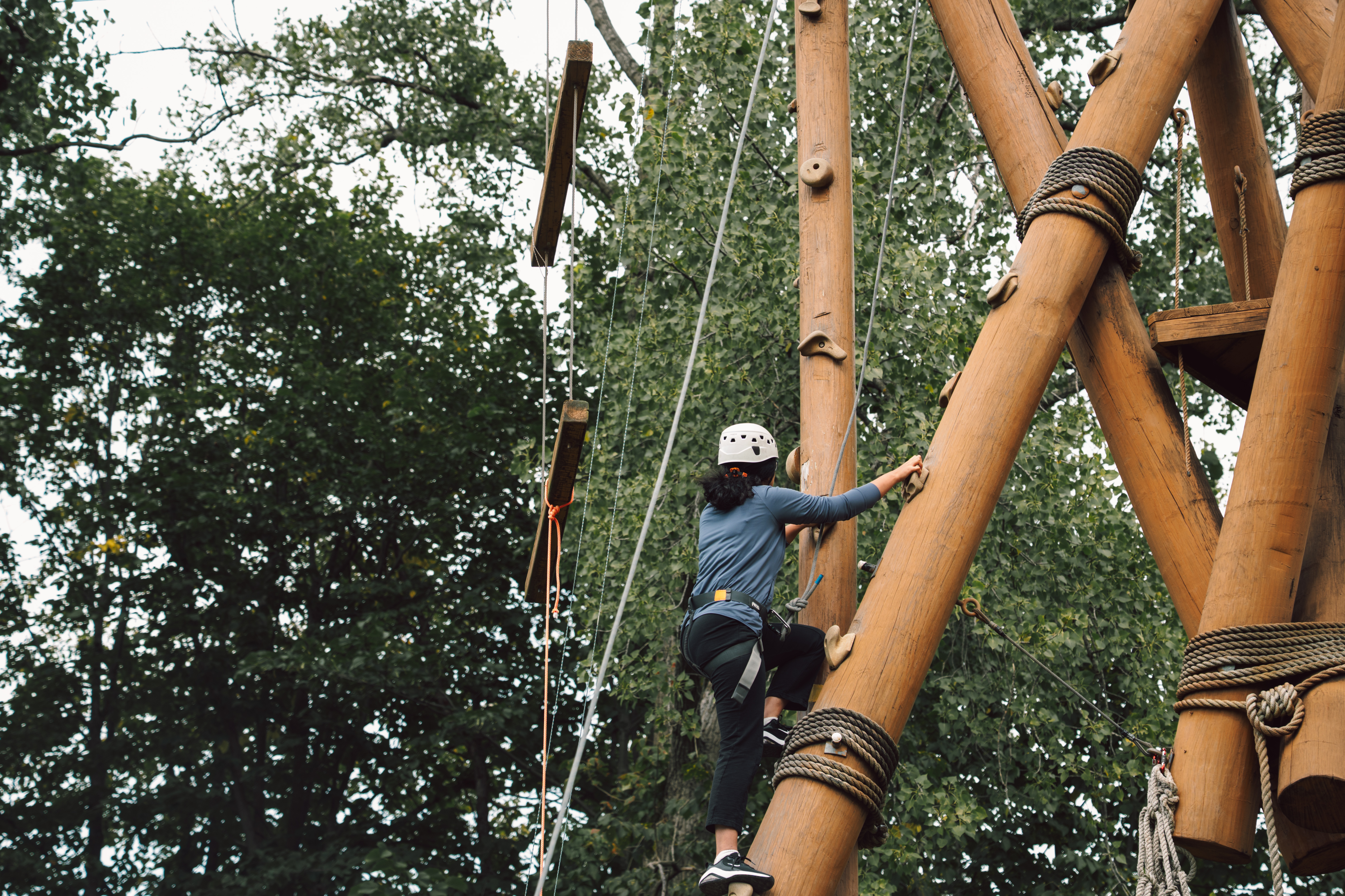 Person wearing a helmet and safety harness climbs a tall wooden ropes course structure outdoors, with trees in the background and suspended planks visible above.