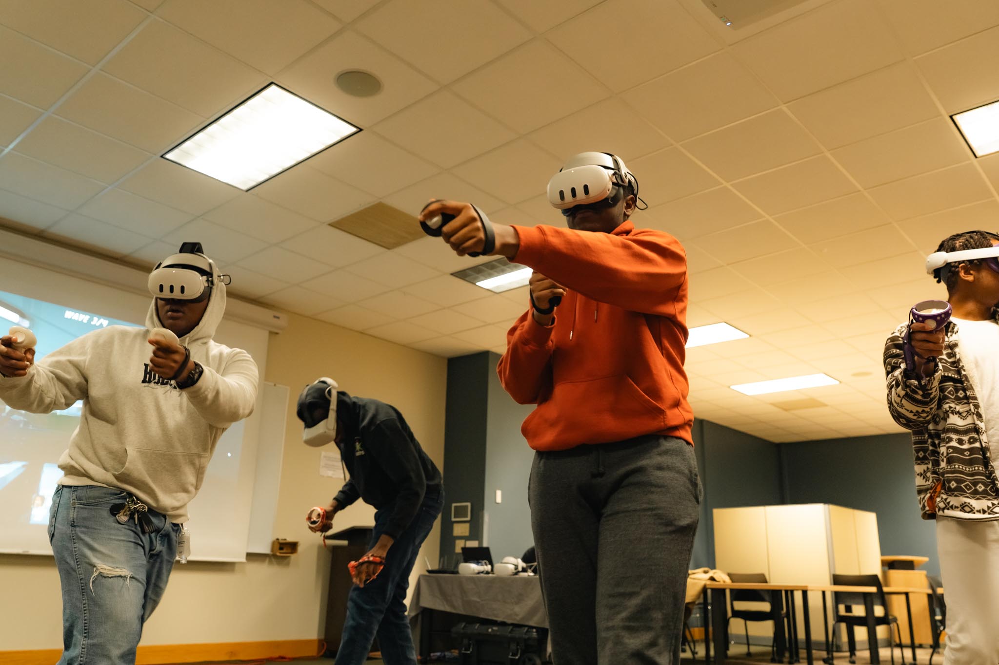 Four people wearing virtual reality headsets and holding controllers participate in an energetic VR activity, with one person in an orange hoodie appearing to punch forward.