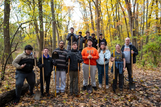 thirteen people stand with yard work tools in a wooded area in the fall.
