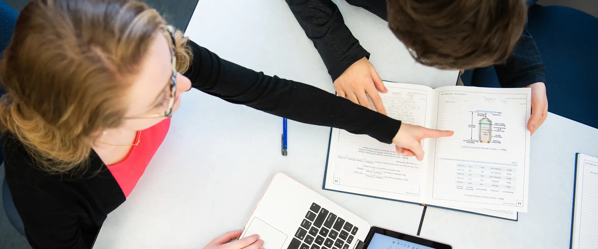 Overhead view of two people sitting at a table looking at a book and laptop.