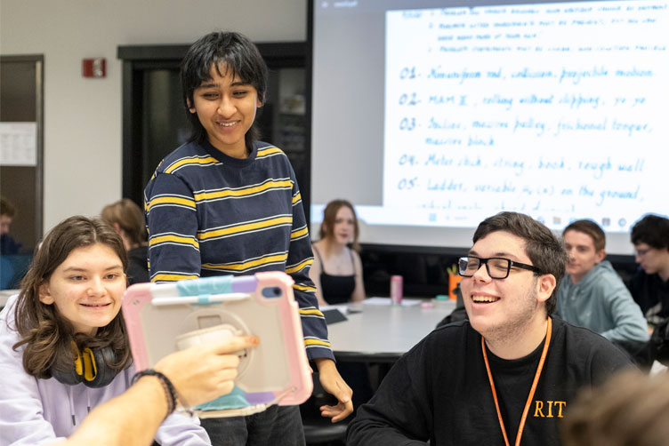 LA is standing behind 2 students working, and looking at the notebook a student sitting opposite direction showing. 