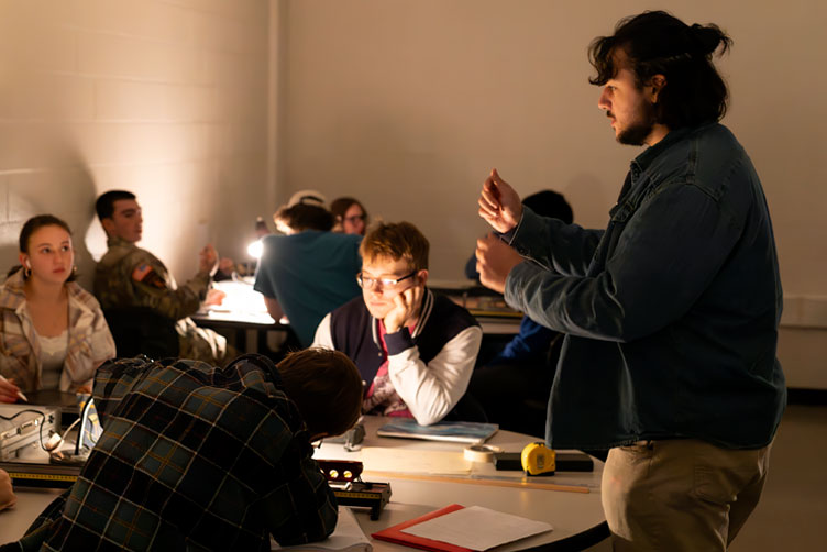 LA is standing over group of students explaining, Physics lab is dark with only table lamp on. 
