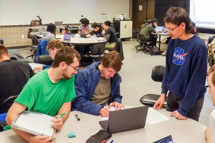Students collaborating in a classroom, with three students in the foreground discussing work at a table with a laptop and papers. Other groups are working at tables in the background.