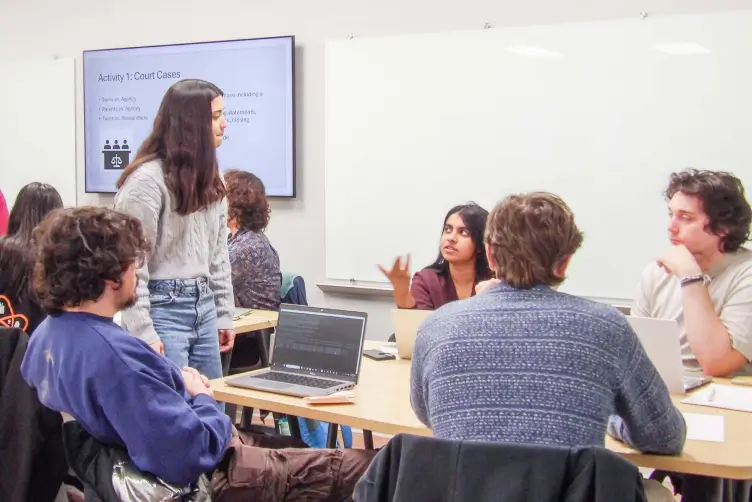 A group of students seated at a classroom table engaged in discussion, while a standing student listens.