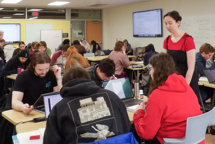 Students working in groups in a busy classroom, with a woman standing and observing one group in the foreground. Laptops and papers cover the tables, and information is displayed on wall monitors.