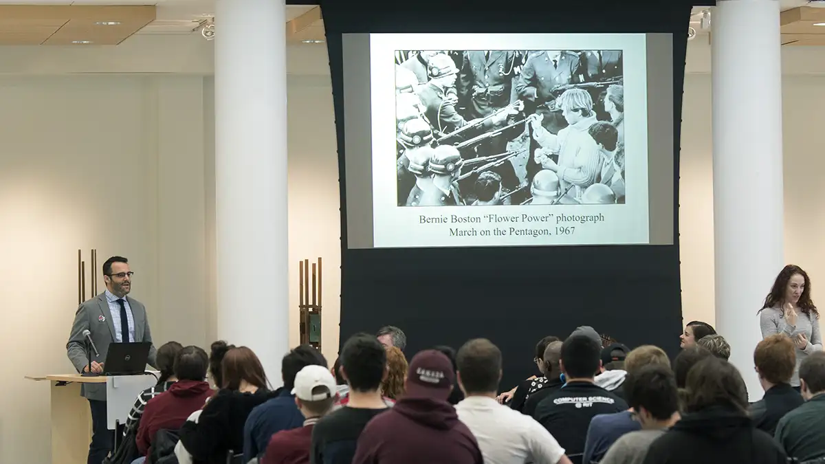 A speaker presents to an audience beside a projected image of Bernie Boston’s 1967 “Flower Power” photograph.