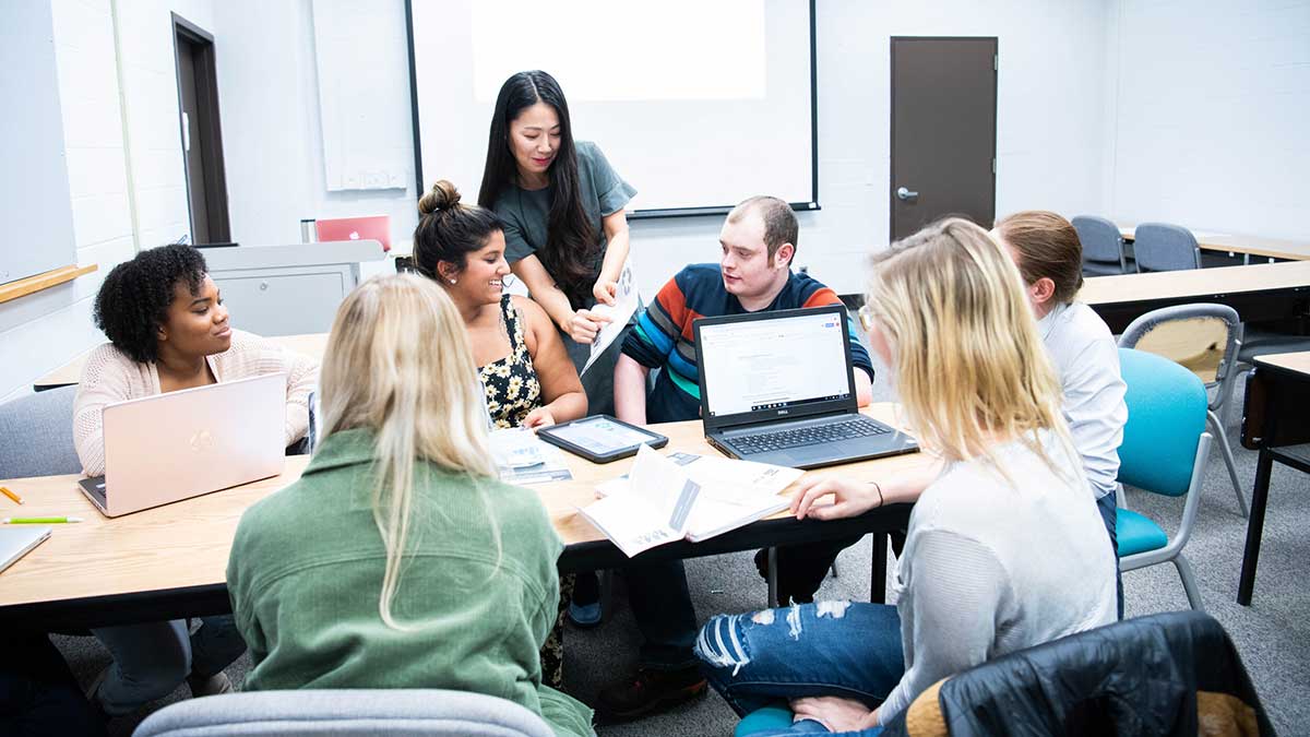Students sit at classroom tables, actively taking notes and listening during a lecture.