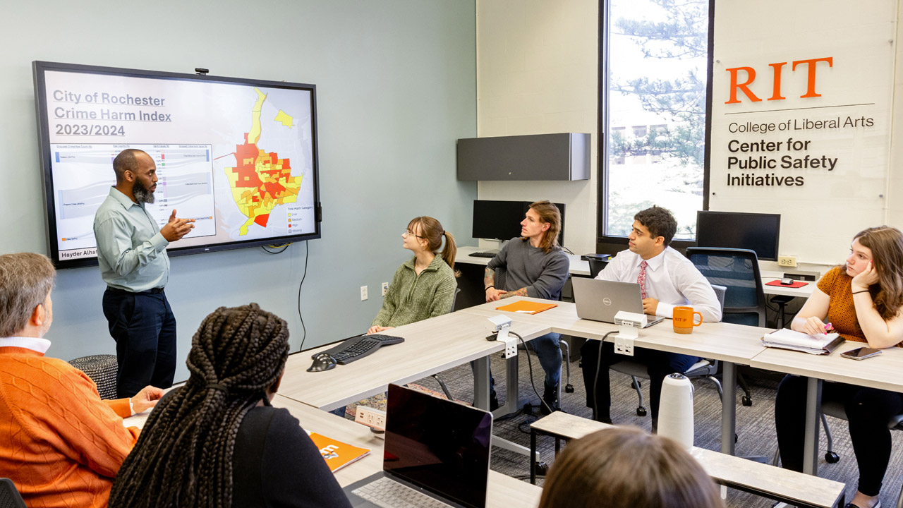 Students listen to a presenter with information about the City of Rochester's Harm Index displayed in the background.