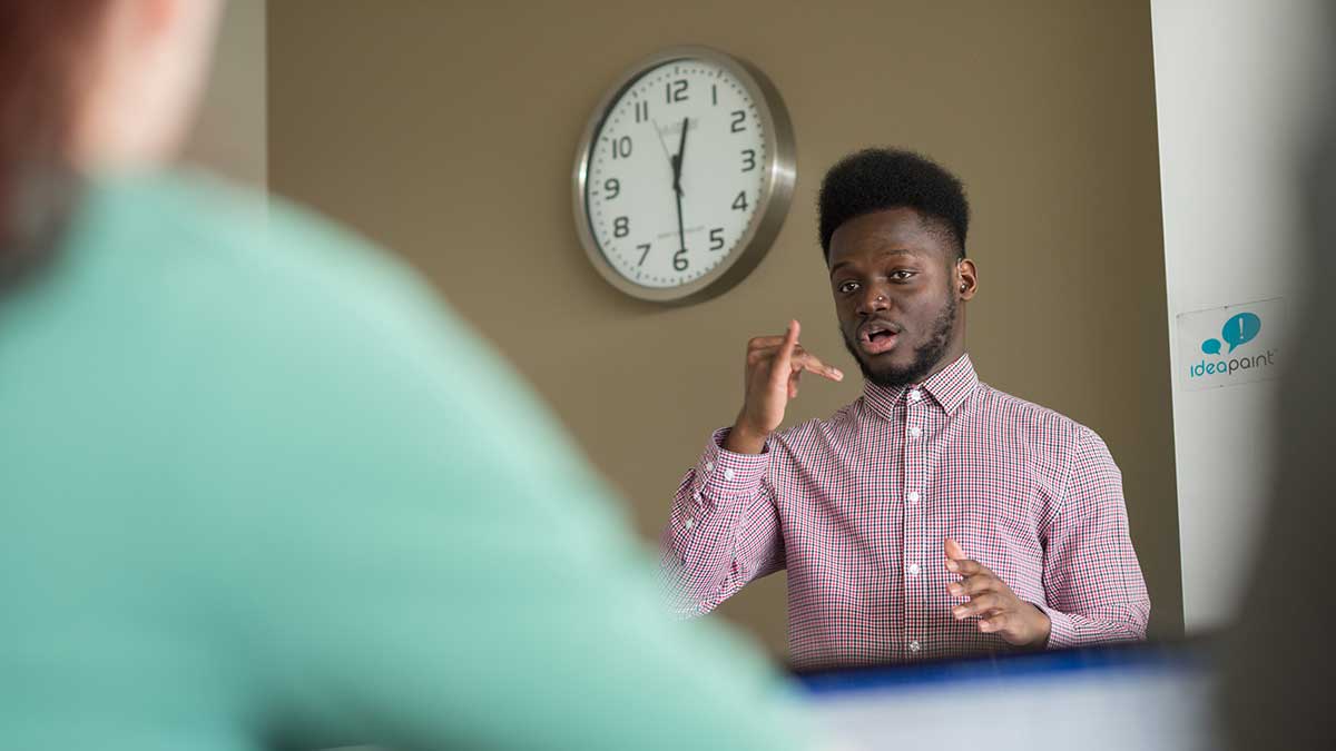 A young man in a checkered shirt using sign language in a classroom setting.