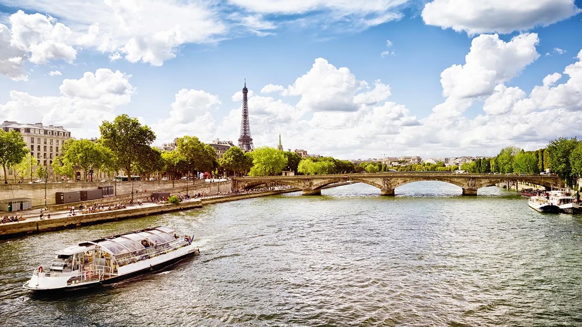 A scenic view of the Eiffel Tower and the Seine River with a boat cruising and people walking along the banks.