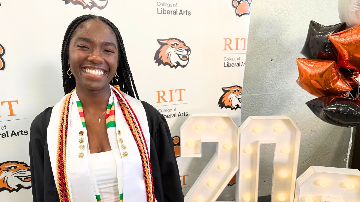 A smiling RIT graduate poses in front of a backdrop with RIT logos and light-up numbers reading 20 24.