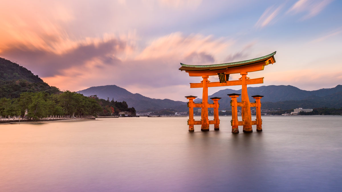 A traditional red torii gate stands in the water with mountains and a vibrant sky in the background.