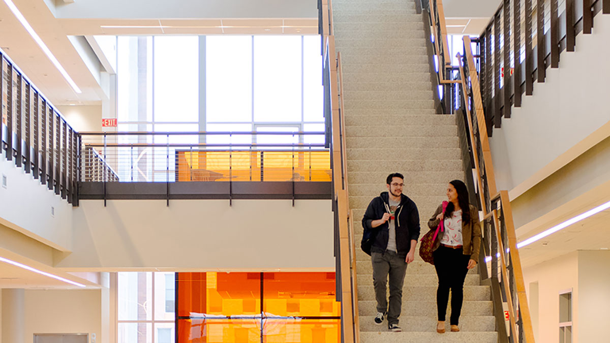 A spacious and brightly lit university atrium with two people walking down a large staircase.
