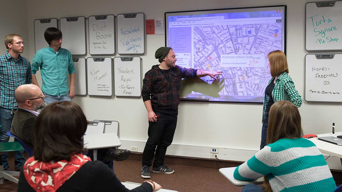 A classroom setting where a student presents a map on a projector screen while others engage in discussion.