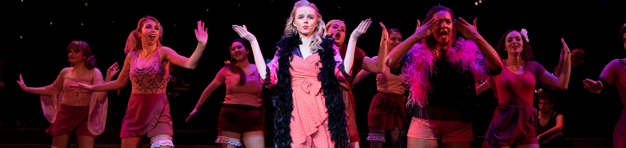 Students on stage under pink lighting in a production of Cabaret. The performer in the middle is more in the spotlight.